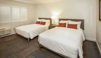 This image shows a hotel room with two neatly made double beds, each with orange pillows, a central nightstand, a lamp, and a large window.