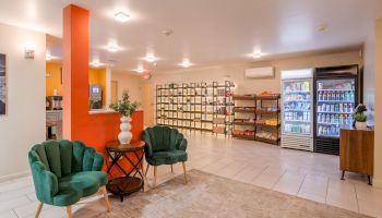 A well-lit lobby with green chairs, snacks on shelves, a fridge with drinks, and a counter beside an orange column.