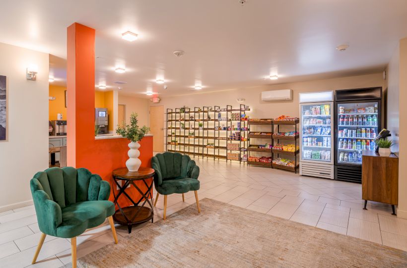 A well-lit lobby with green chairs, snacks on shelves, a fridge with drinks, and a counter beside an orange column.