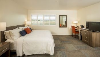 A spacious bedroom with a large bed, desk, chair, TV, and nightstands, featuring a window with shutters and neutral color decor, ending the sentence.