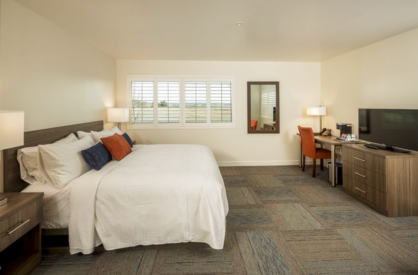 A spacious bedroom with a large bed, desk, chair, TV, and nightstands, featuring a window with shutters and neutral color decor, ending the sentence.