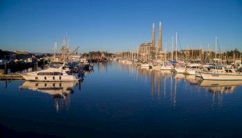 A calm marina with boats docked in still water reflecting the clear blue sky and nearby structures, including a facility with tall chimneys.