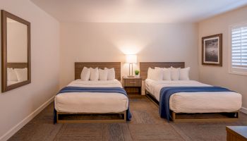 This image shows a hotel room with two double beds, a nightstand with a lamp, a mirror, and a framed picture on the wall.