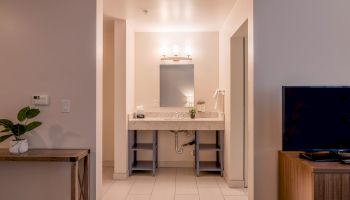 A clean bathroom with a mirror, lighting, sink, and a neatly organized vanity. A TV and wooden console are visible on the right side.