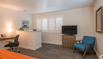 A cozy room with a desk, chair, TV, blue armchair, lamp, and shutters on the window. Modern decor with an orange bedspread.