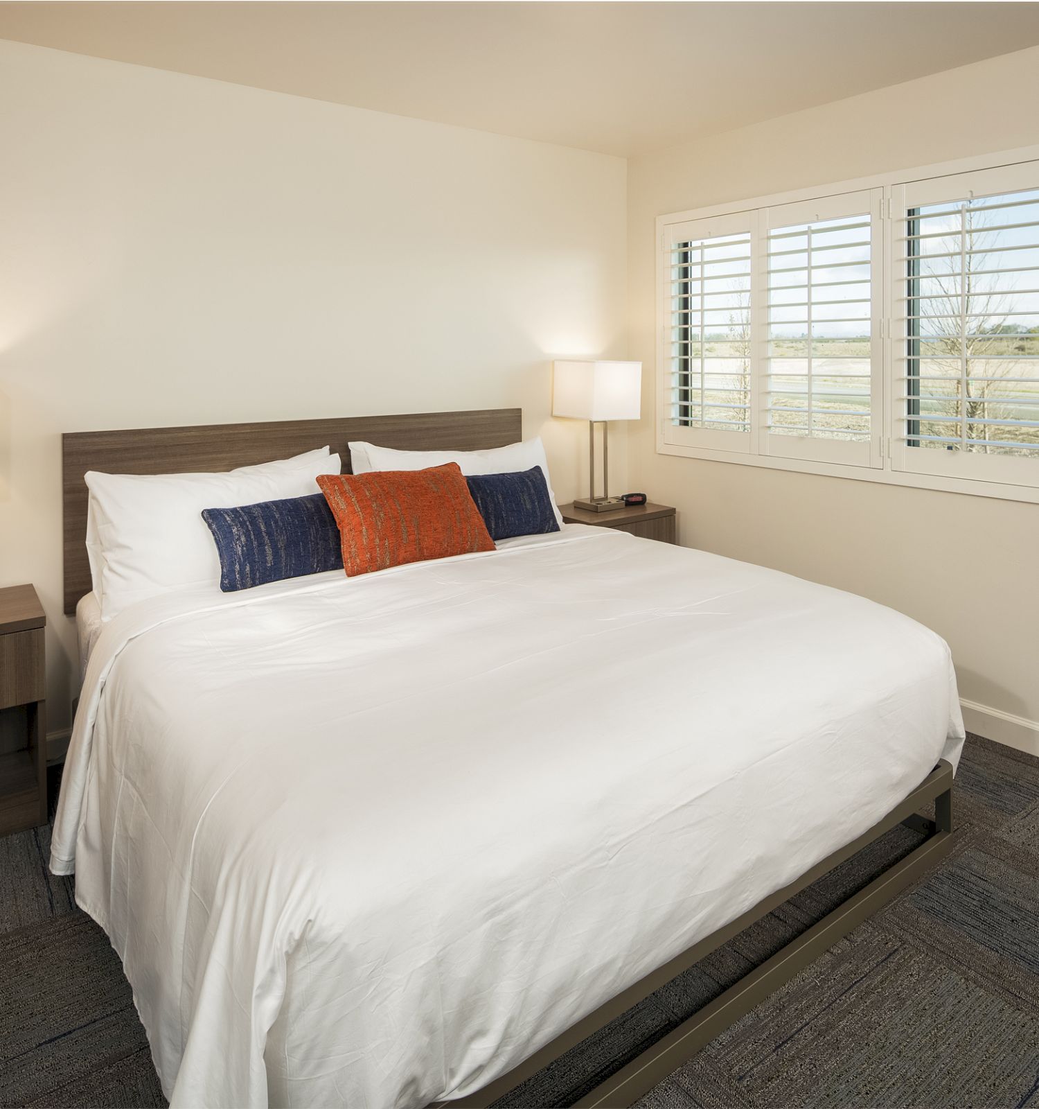 A neatly arranged bedroom with a large bed, white bedding, and colorful pillows. There are two bedside tables with lamps, and a window with shutters.