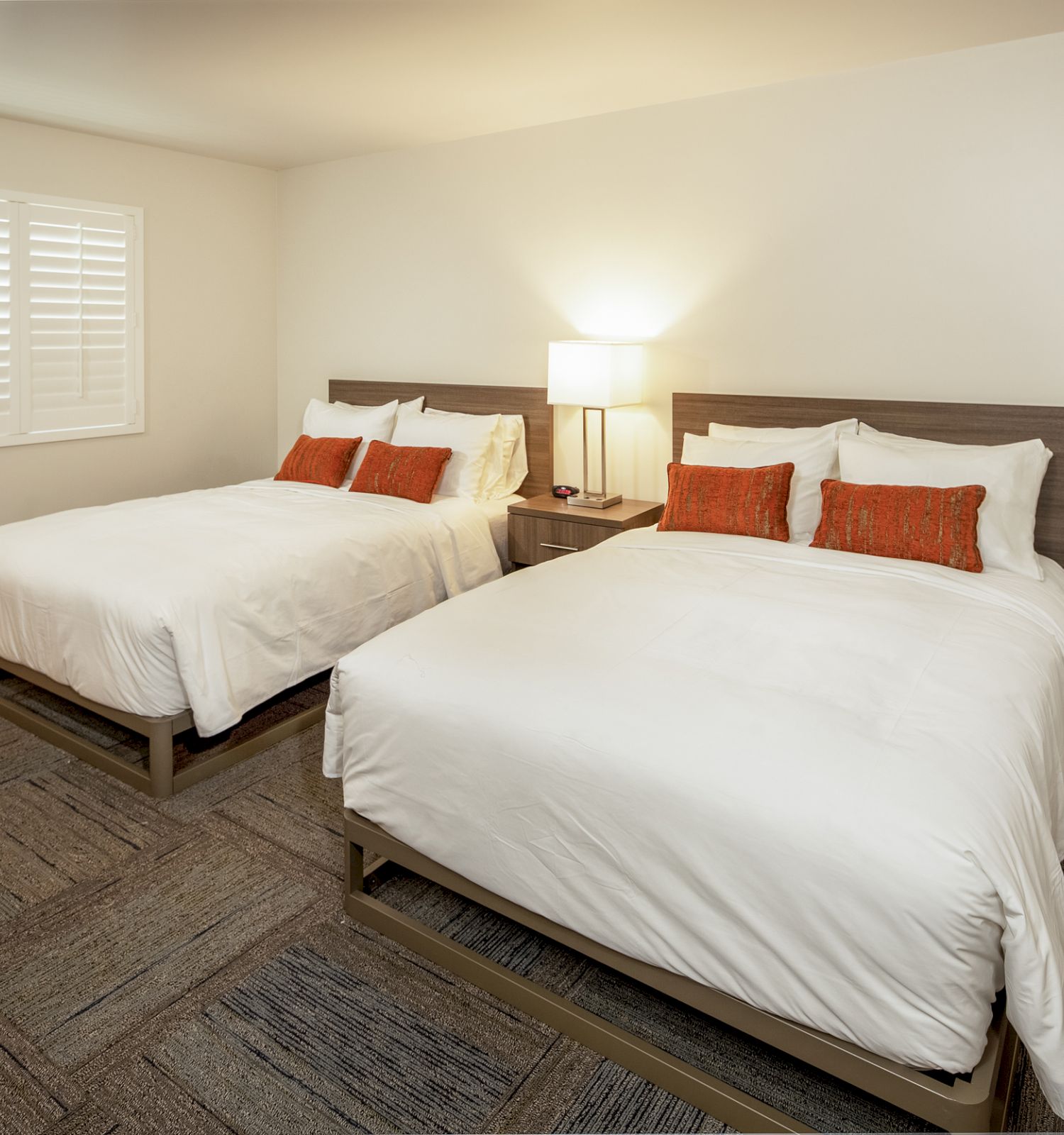 A minimalistic bedroom has two white beds with red pillows, a wooden nightstand with a lamp, white walls, and a window with shutters.