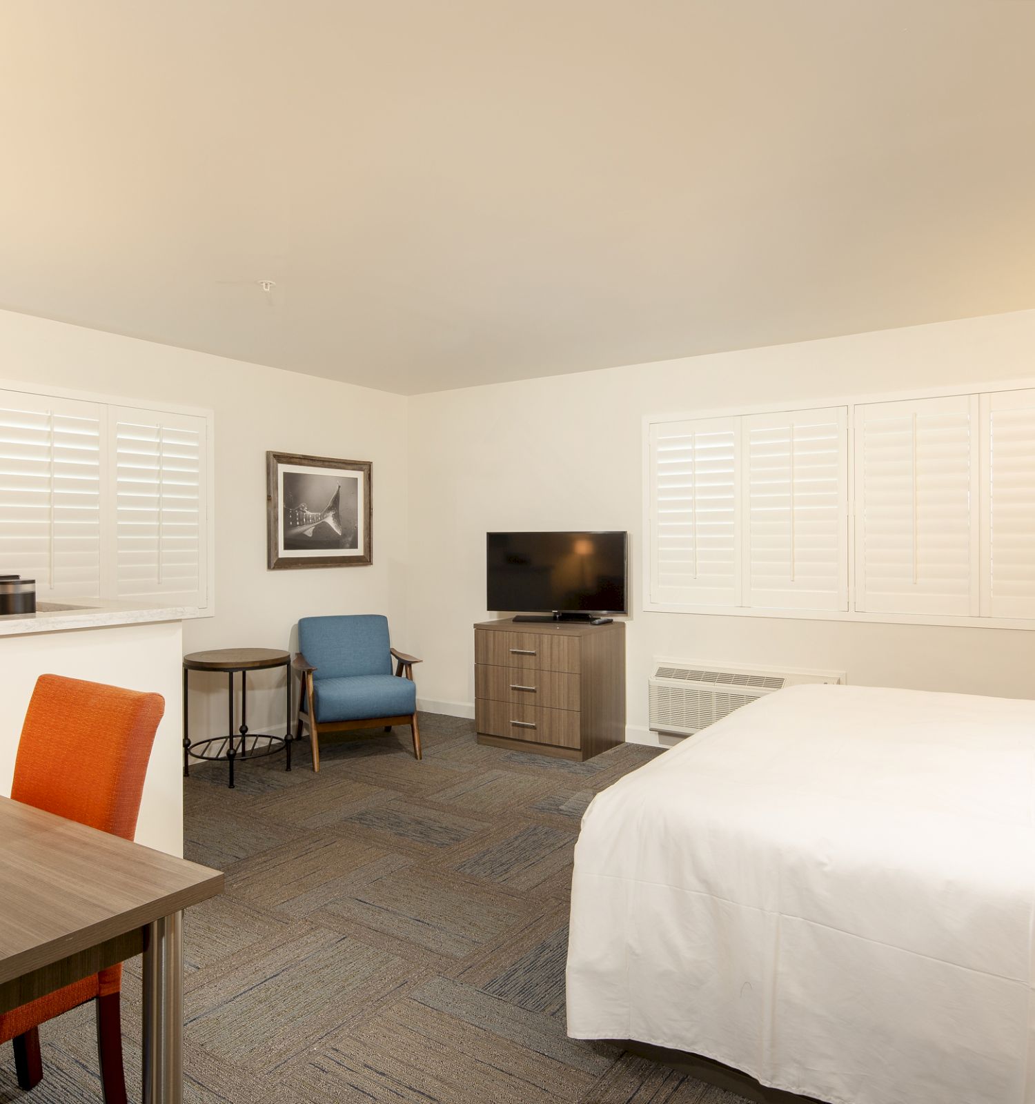 A spacious hotel room with a neatly made bed, desk, chairs, television, and artwork on the wall. The windows have white shutters, and there's soft lighting.