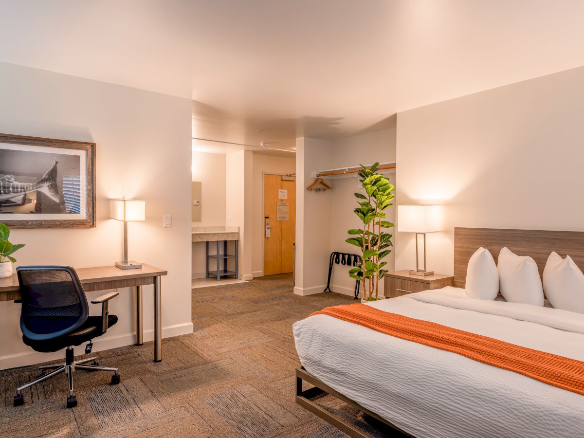 A modern hotel room with a bed, desk, chair, plants, and artwork; cozy lighting and open closet space complete the serene setting.