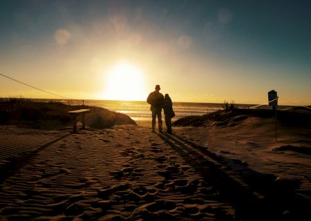 A couple stands on a beach, silhouetted by the setting sun. The scene includes sand, footprints, and a bench, creating a peaceful and romantic atmosphere.