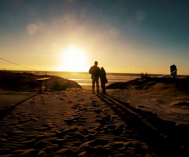 A couple stands on a beach, silhouetted by the setting sun. The scene includes sand, footprints, and a bench, creating a peaceful and romantic atmosphere.