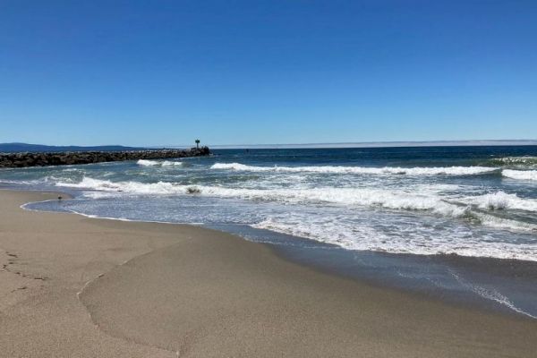 A serene beach scene with clear blue skies, gentle waves washing onto the sandy shore, and a distant rock formation under a cloudless horizon.