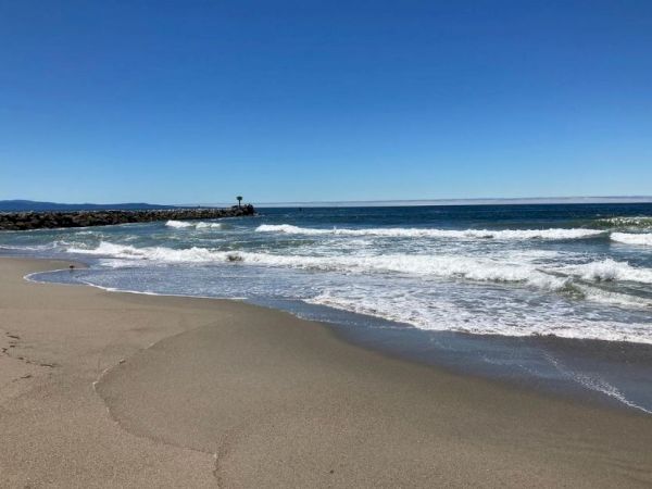 A serene beach scene with clear blue skies, gentle waves washing onto the sandy shore, and a distant rock formation under a cloudless horizon.