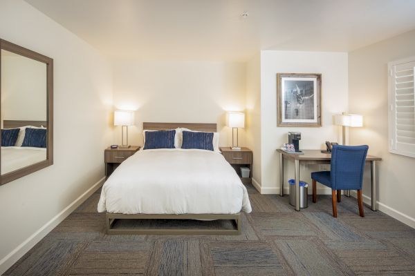 A hotel room with a neatly made bed, two nightstands, lamps, a desk with a chair, a coffee maker, and a large wall mirror.