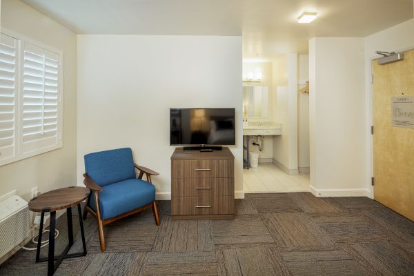 The image shows a minimalistic room with a blue chair, small table, TV on a stand, carpeted floor, and a bathroom in the background, ending the sentence.