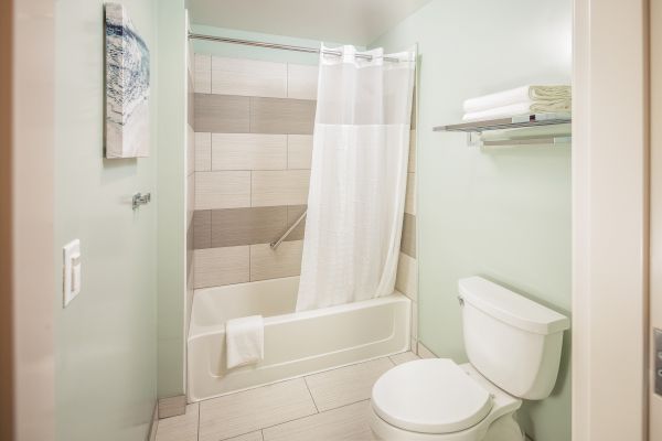 The image shows a clean, modern bathroom with a bathtub, shower curtain, toilet, towel rack, and wall art. The room has light-colored tiles and walls.