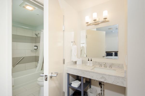 A clean, modern bathroom with a bathtub, shower, sink, and mirror under bright lights. The sink area has toiletries and a towel rack underneath.