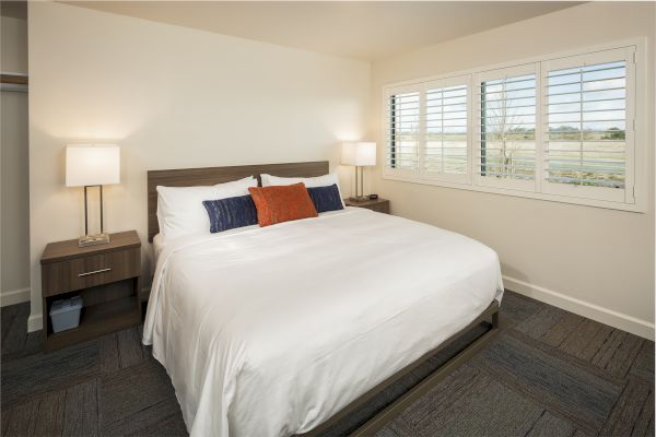 A modern bedroom with a large bed, white bedding, and colorful accent pillows. Two bedside tables with lamps and a window with shutters are present.