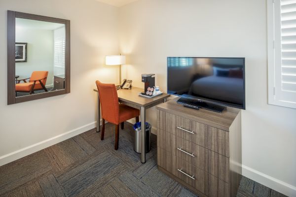 The image shows a room with a desk, orange chair, lamp, phone, TV, drawer unit, and a mirror on the wall.