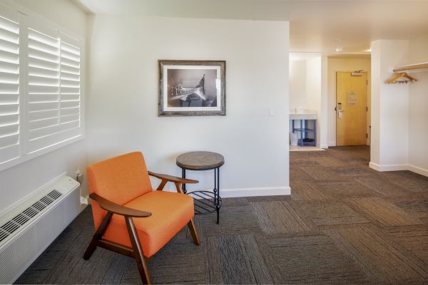 A room with an orange chair, round table, wall picture, closet, and door. White walls and carpeted floor with window blinds.