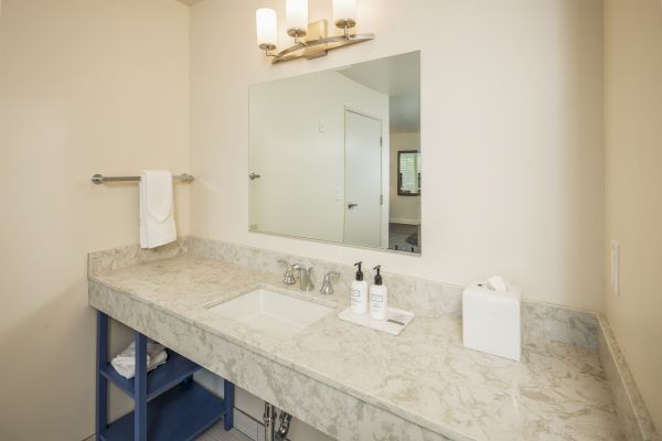 This image shows a bathroom with a marble countertop, a rectangular mirror, a sink, toiletries, a tissue box, a towel on a rod, and vanity lights.