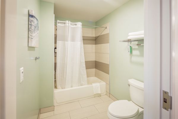 A clean, modern bathroom with a bathtub and shower curtain, toilet, towel shelf, and wall art, featuring light green walls and tiled flooring.