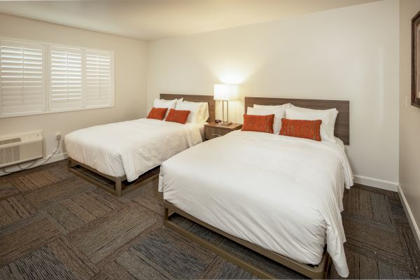 A hotel room with two double beds, white bedding, orange accent pillows, a bedside table, a lamp, a window with shutters, and carpeted floor.