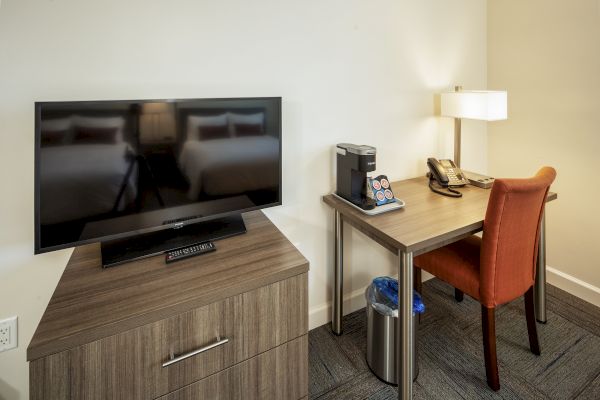 A room with a TV on a dresser, a desk with a lamp, coffee maker, and chair, and a trash bin underneath the desk ending the sentence.