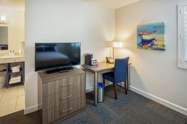 A hotel room workspace with a TV on a dresser, a desk with a lamp, phone, coffee maker, and a blue chair, and a painting on the wall.