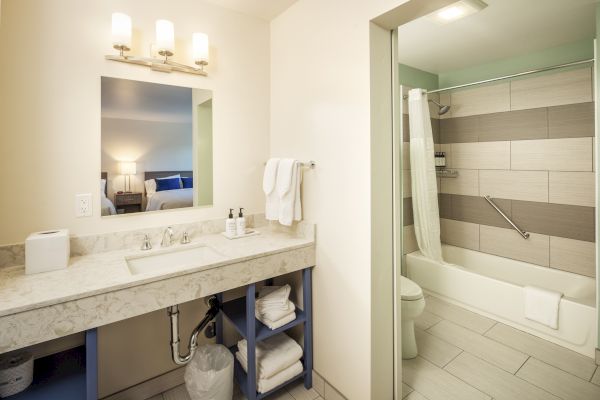 Modern bathroom with a large mirror, sink, towels, and toiletries. In the background, there is a shower-tub combo with a shower curtain.