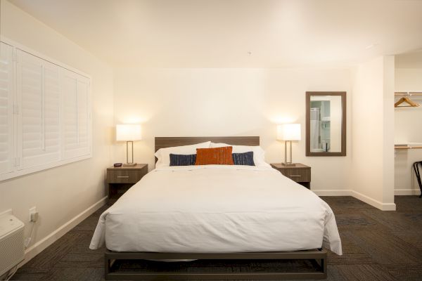 A neatly made bed in a well-lit room with two bedside tables, lamps, a wall mirror, and a small closet area with hangers.