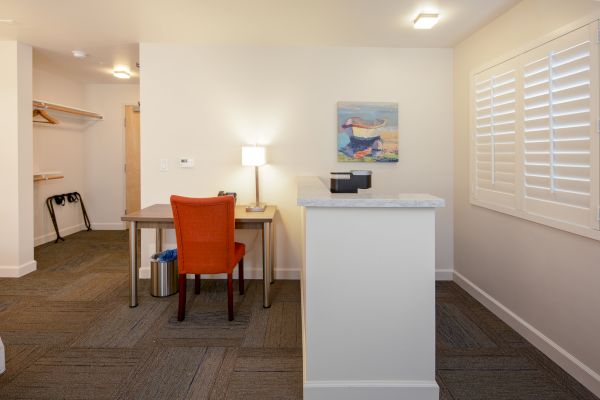 A room with a desk, orange chair, lamp, artwork on the wall, open closet with hangers, and window with shutters.