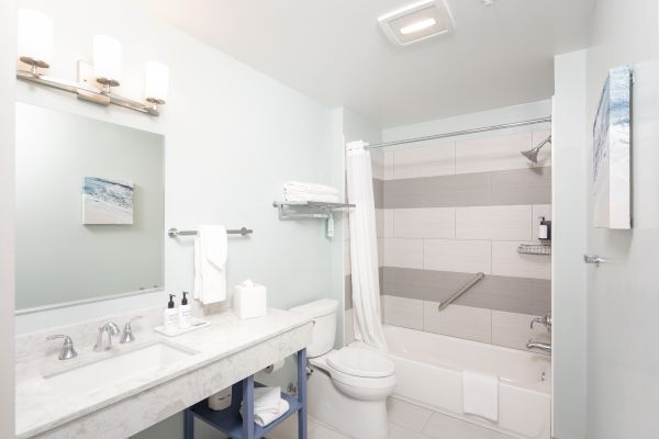 This image shows a modern bathroom with a sink, mirror, towel racks, toiletries, a bathtub with a curtain, and a toilet, in neutral tones.