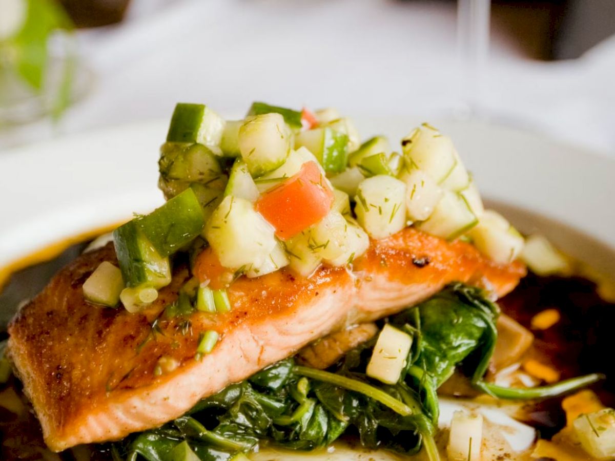 Grilled salmon topped with a cucumber and tomato salad on a bed of sautéed spinach, served with sauce.