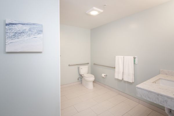 A clean, modern bathroom with a toilet, white towels on a rack, a painting on the wall, and a sink with a marble countertop.