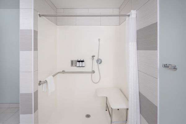 The image shows a clean, modern shower with a bench, handrails, a showerhead, bottles, and a towel hanging on a rail, enclosed by a light-colored curtain.