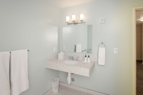 A clean bathroom with a countertop sink, mirror, lighting, towel rack with towels, and a waste bin is visible. The door is partially open.