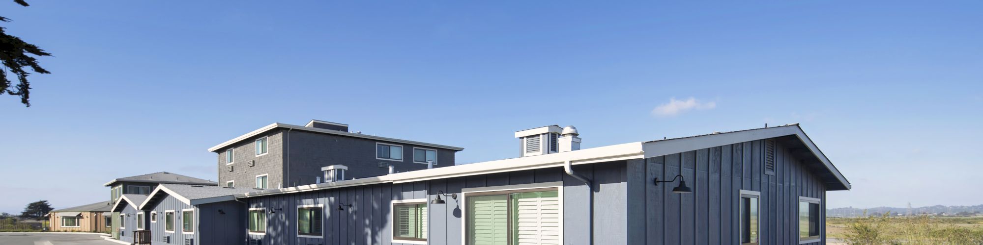 The image shows several modern, single-story buildings in a coastal setting with a clear blue sky in the background.