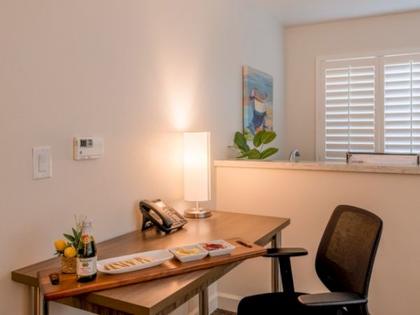 A cozy workspace with a wooden desk, lamp, office chair, phone, snacks, and a drink, accented by art and a plant.