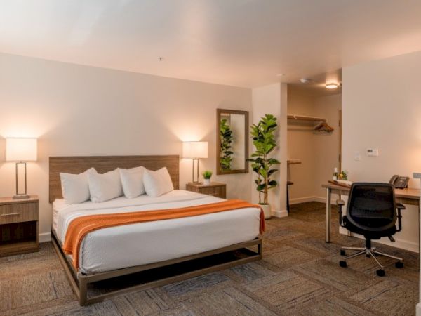 A cozy hotel room with a king-size bed, orange accents, a desk with a chair, and a small closet area, warmly lit by lamps.