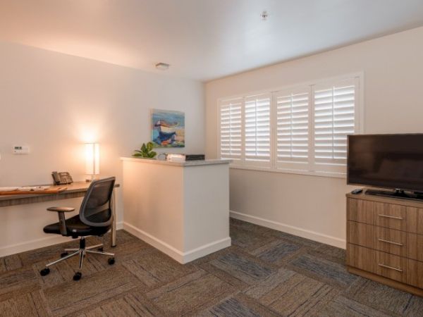 A modern office with a desk, chair, lamp, phone, TV, and window shutters. Decor includes a painting and indoor plants for a cozy atmosphere.