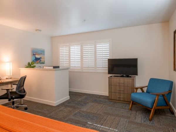 The image shows a modern room with a TV, blue chair, desk with chair, and artwork. There's a bed with an orange blanket in view.