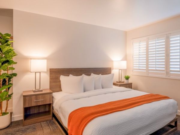 A neatly arranged bedroom with a bed, orange throw, two lamps, plants, and a window with shutters, creating a cozy atmosphere.