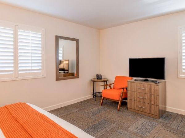 A bedroom with an orange chair, wooden dresser, TV, mirror, and bed with orange accents. White shutters cover the windows.