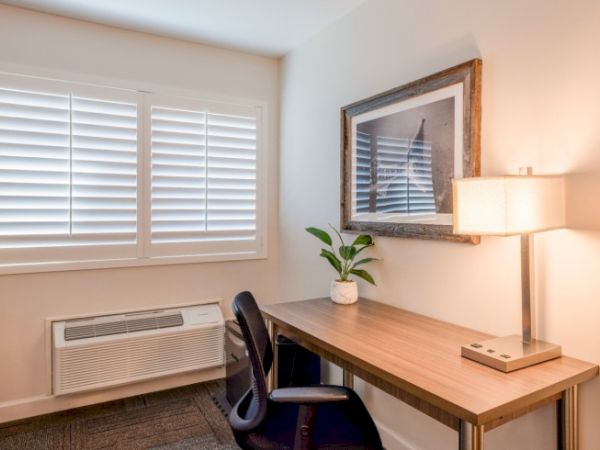 A small office space with a desk, chair, lamp, framed picture, and potted plant. Window with shutters, and an air conditioning unit below it.