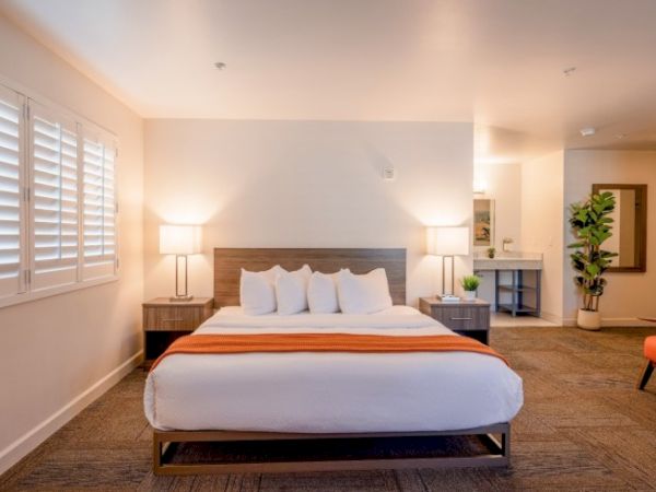 A well-lit bedroom with a large bed, orange throw, bedside tables, lamps, and a plant, creating a cozy and inviting atmosphere.