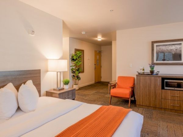 A cozy hotel room with a bed, orange chair, lamp, dresser, microwave, and decor. A doorway leads outside. Walls are neutral colored.