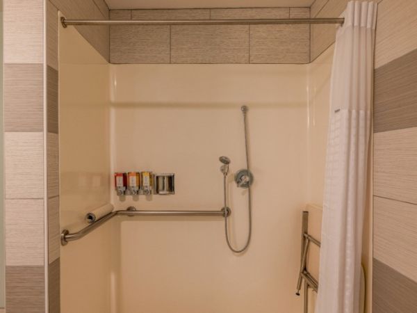 The image shows an accessible shower with a wall-mounted showerhead, grab bars, shower curtain, and toiletries on the wall.