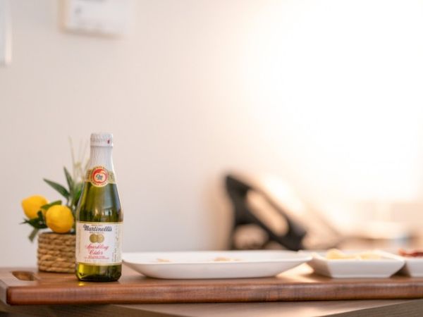 A bottle of sparkling juice sits on a wooden tray with a potted plant and empty dishes, against a softly lit background.