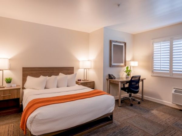 A modern hotel room with a bed, desk, and lamps, featuring an orange throw blanket and a window with blinds.